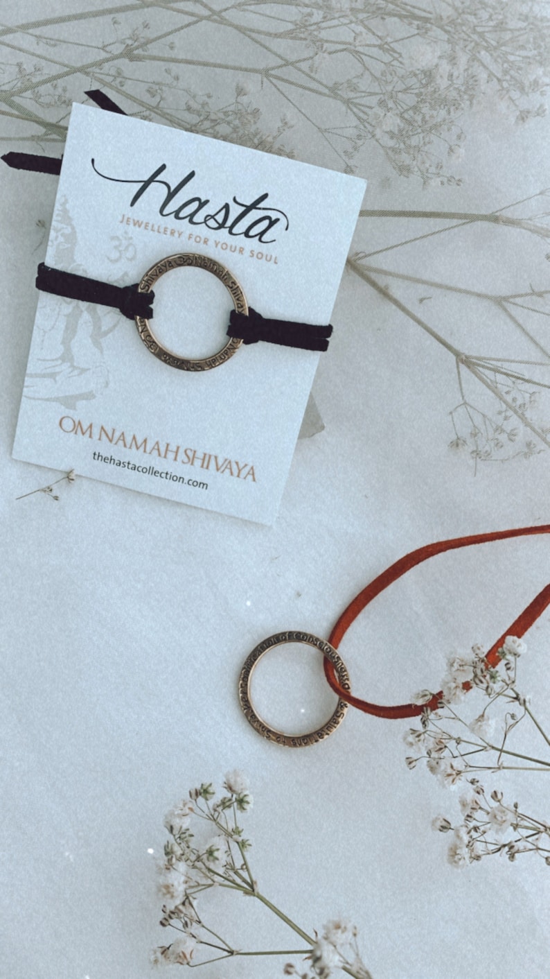 May include: A black cord bracelet with a gold-toned circular charm inscribed with the phrase "Om Namah Shivaya". The bracelet is displayed on a white card with the brand name "Hasta" and the tagline "Jewellery for your soul".