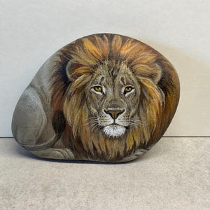 May include: Hand-painted rock art depicting a majestic lion. The lion's face is detailed with a brown mane, yellow eyes, and a white muzzle. The rock is oval-shaped and rests on a light gray surface.