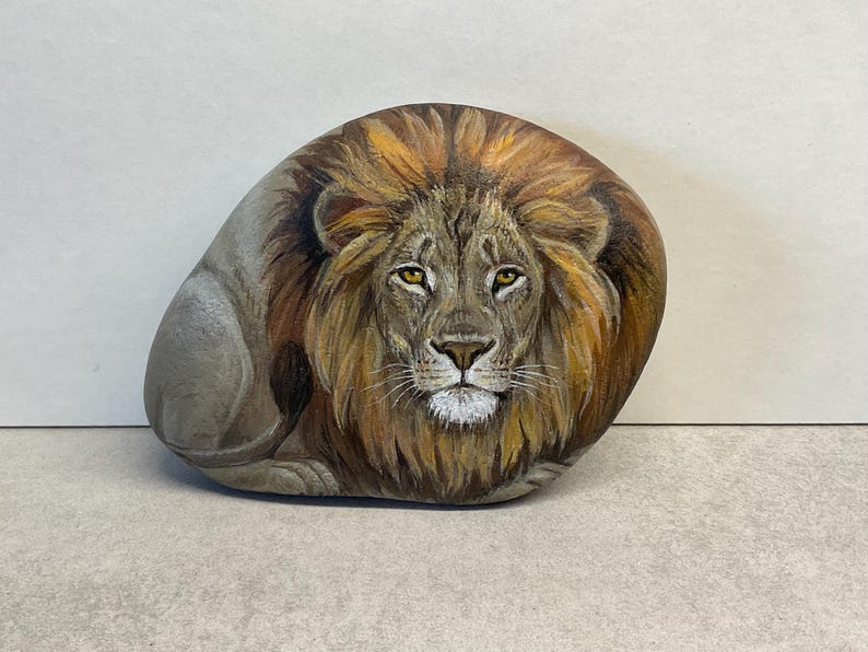 May include: Hand-painted rock art depicting a majestic lion. The lion's face is detailed with a brown mane, yellow eyes, and a white muzzle. The rock is oval-shaped and rests on a light gray surface.