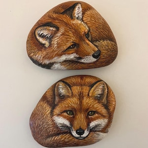 Pode incluir: Duas pedras pintadas à mão com retratos detalhados de raposas. As raposas têm pelo laranja e castanho, detalhes brancos e olhos verdes. As pedras são lisas e ovais, perfeitas para decoração de casa.