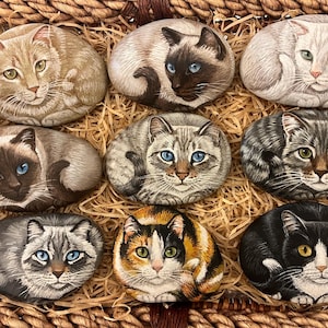May include: A collection of nine hand-painted cat rocks displayed in a woven basket. The rocks showcase diverse cat breeds, including tabby and Siamese, with detailed facial features and fur patterns. The cats are painted in various colors and rest on a bed of straw, creating a charming display.