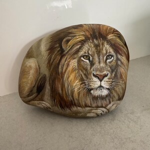 May include: A hand-painted rock features a detailed depiction of a lion's face and mane. The artwork uses shades of brown, tan, and gold to create a realistic and textured appearance. The lion is painted on a smooth, oval-shaped rock.