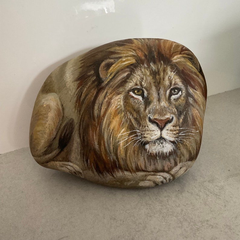 May include: A hand-painted rock features a detailed depiction of a lion's face and mane. The artwork uses shades of brown, tan, and gold to create a realistic and textured appearance. The lion is painted on a smooth, oval-shaped rock.