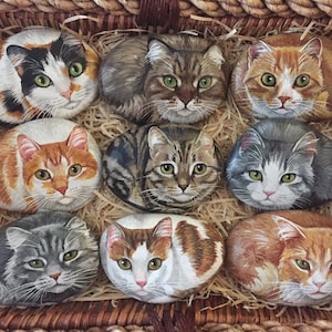 Puede incluir: Una cesta de mimbre llena de piedras pintadas con temática de gatos. Las piedras están pintadas con representaciones realistas de gatos en varias poses y colores, incluyendo atigrado, calico y jengibre.