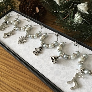 May include: A set of six silver-toned wine glass charms arranged in a rectangular box. Each circular charm features alternating light blue and white beads, small rhinestone spacers, and a unique holiday-themed metal pendant including a stocking, snowflake, gingerbread man, tree, and reindeer.