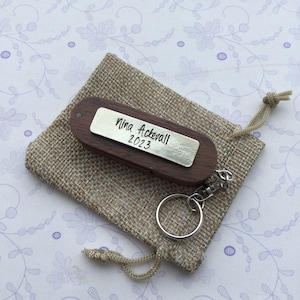 May include: A wooden keychain with a silver nameplate engraved with "Nina Ackevall 2023" rests on a small burlap bag. The keychain has a silver keyring and clasp. The bag is tied with a drawstring.