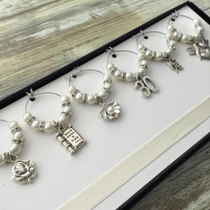 May include: A set of six silver-toned wine glass charms featuring white faux pearls and clear rhinestones. Each charm has a unique dangling pendant including a rose, a book with the text Once Upon a Time, a seashell, the number 30, the word Love, and a bird.
