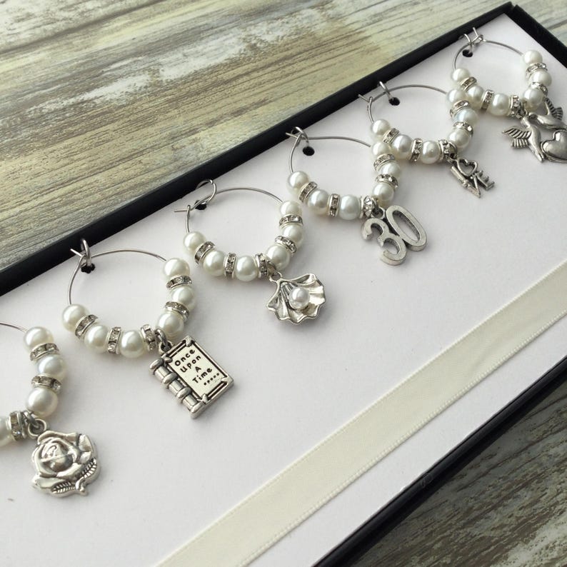 May include: A set of six silver-toned wine glass charms featuring white faux pearls and clear rhinestones. Each charm has a unique dangling pendant including a rose, a book with the text Once Upon a Time, a seashell, the number 30, the word Love, and a bird.