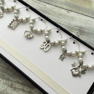 May include: A set of six silver-toned wine glass charms featuring white faux pearls and clear rhinestones. Each charm includes a unique metal pendant, including a book with the text Once Upon a Time, a seashell, the number 30, the word LOVE, and two birds.