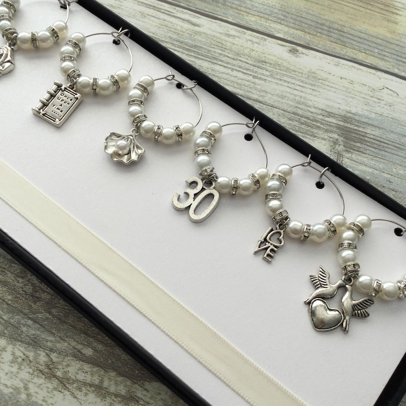 May include: A set of six silver-toned wine glass charms featuring white faux pearls and clear rhinestones. Each charm includes a unique metal pendant, including a book with the text Once Upon a Time, a seashell, the number 30, the word LOVE, and two birds.