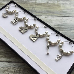 May include: A set of six silver-toned wine glass charms featuring white faux pearls and clear rhinestones. The charms include individual pendants shaped like a shell, the number 30, hearts inscribed with Yvonne and Ward, a wine glass, and a teddy bear.