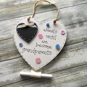 May include: A white heart-shaped wooden sign with the text "weeks until we become grandparents." It features a small black chalkboard heart, pink and blue buttons, and a ribbon. A rolled scroll is attached.