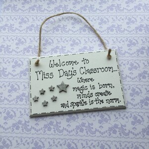 May include: White wooden sign with black text that reads "Welcome to Miss Day's Classroom... Where magic is born, minds create and sparkle is the norm." The sign has silver glitter stars.