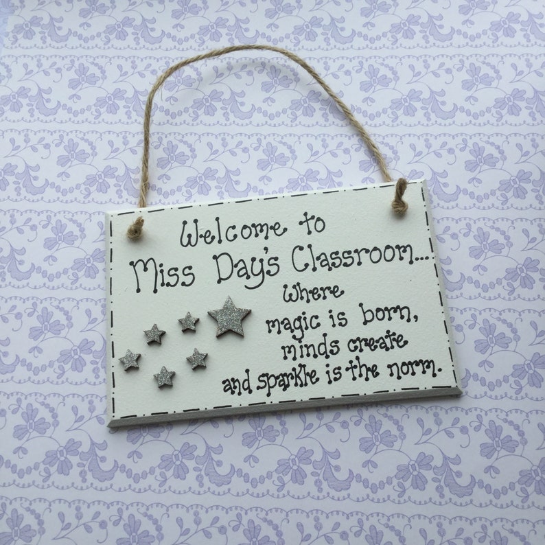 May include: White wooden sign with black text that reads "Welcome to Miss Day's Classroom... Where magic is born, minds create and sparkle is the norm." The sign has silver glitter stars.
