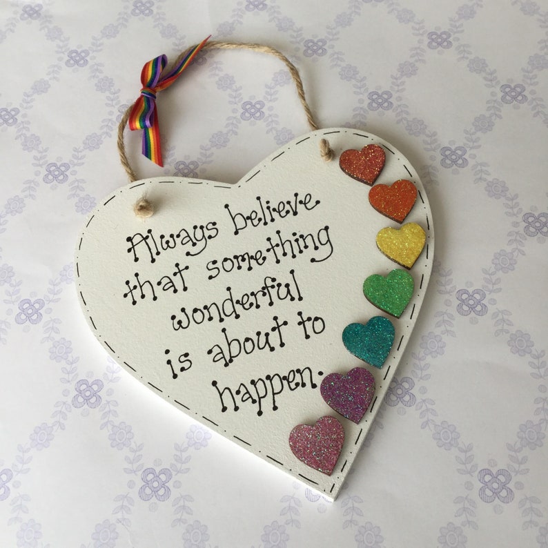 May include: White wooden heart-shaped sign with the text "Always believe that something wonderful is about to happen."  The sign is decorated with a rainbow of glitter hearts.