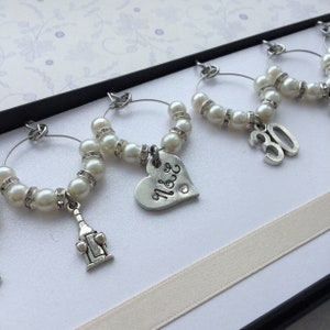 May include: A set of silver-toned wine glass charms featuring white faux pearls and clear rhinestones. The charms include a silver bottle-shaped pendant, a heart-shaped charm engraved with N&E, and a silver number 30 charm, all on circular wire hoops.