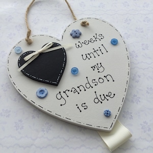 May include: A white heart-shaped wooden sign with the text "weeks until my grandson is due." It features a black heart-shaped chalkboard, blue buttons, and a cream ribbon. A twine loop is at the top for hanging.