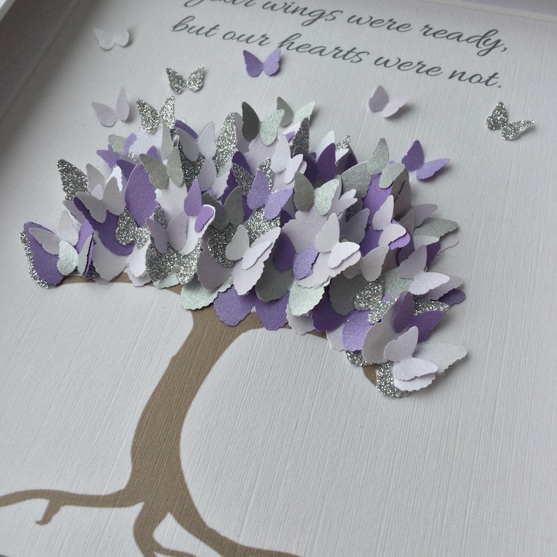 May include: A framed artwork featuring a tree with a brown trunk and branches, adorned with numerous paper butterflies in shades of purple, silver, and white. The butterflies appear to be 3D, adding depth to the piece. The background is white with text.