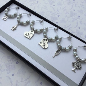 May include: A set of six silver-toned wine glass charms arranged in a black box. Each circular charm features white faux pearls and clear rhinestones, with unique dangling pendants including the number 25, a bottle, a heart with R&H, a heart with 18.07.97, a champagne flute, and a tree.