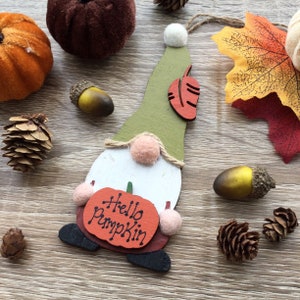 Puede incluir: Adorno de gnomo de madera con un sombrero verde, barba blanca y una calabaza roja que dice "Hello Pumpkin". El gnomo está sosteniendo la calabaza en sus manos. El adorno está rodeado de decoraciones de otoño, incluyendo bellotas, piñas y una hoja de otoño.