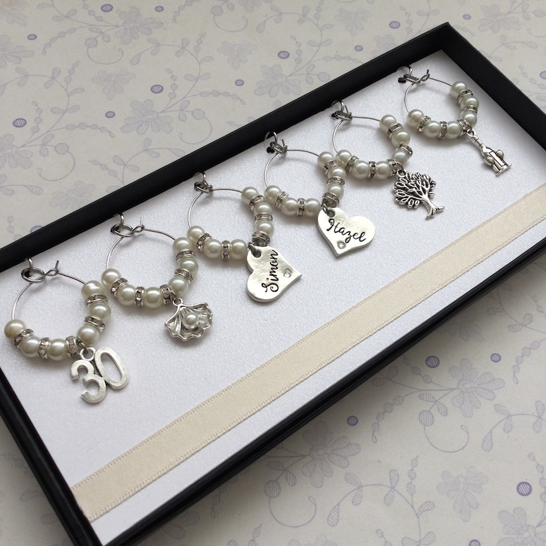 May include: A set of six silver-toned wine glass charms arranged in a black box. Each charm features a circular wire hoop decorated with white faux pearls and clear crystals, accented by unique silver charms including the number 30, a shell, hearts with the names Simon and Hazel, a tree, and a small figure.