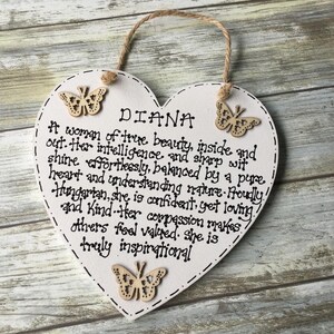 Puede incluir: Placa de madera blanca en forma de coraz&oacute;n con el nombre "DIANA" y un poema. La placa tiene tres adornos de mariposa de madera y un lazo de cuerda para colgar. El texto es negro.