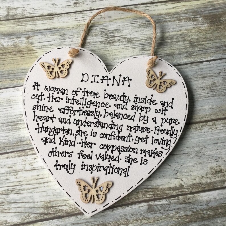 Puede incluir: Placa de madera blanca en forma de coraz&oacute;n con el nombre "DIANA" y un poema. La placa tiene tres adornos de mariposa de madera y un lazo de cuerda para colgar. El texto es negro.