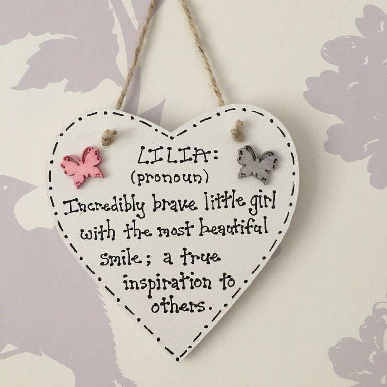 Puede incluir: Un letrero de madera en forma de coraz&oacute;n blanco con el nombre "LILIA" y una descripci&oacute;n. El letrero tiene un borde punteado, adornos de mariposa y una cuerda de yute para colgar. El texto dice "Incredibly brave little girl..."