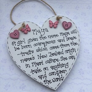 Puede incluir: Placa de madera en forma de coraz&oacute;n blanco con el nombre "Maya" escrito en negro. Decorada con mariposas y corazones rosas. Una cuerda de yute est&aacute; unida para colgarla. El texto describe los rasgos de Maya.