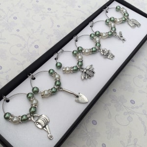 Peut inclure: Ensemble de six charmes pour verres à vin en perles vertes et blanches avec des breloques argentées en forme d'arrosoir, de pelle, d'abeille, de pot de fleurs, d'oiseau et de gant de jardinage.