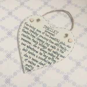 Puede incluir: Placa de madera en forma de coraz&oacute;n, color crema, con el nombre "SHELLEY" y una descripci&oacute;n manuscrita de sus intereses. Cuenta con una cuerda de yute para colgar y peque&ntilde;os corazones de madera.