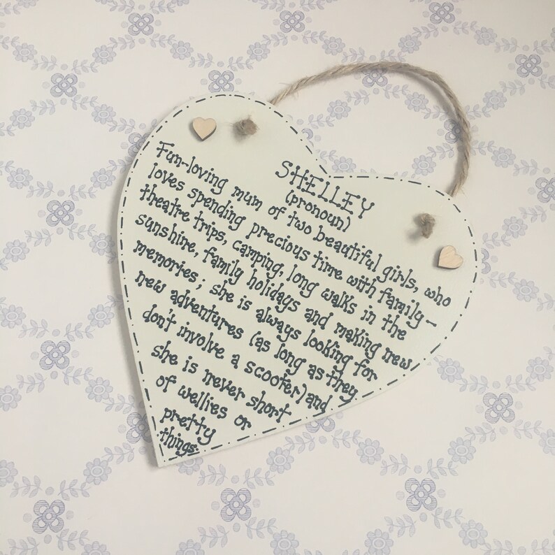 Puede incluir: Placa de madera en forma de coraz&oacute;n, color crema, con el nombre "SHELLEY" y una descripci&oacute;n manuscrita de sus intereses. Cuenta con una cuerda de yute para colgar y peque&ntilde;os corazones de madera.
