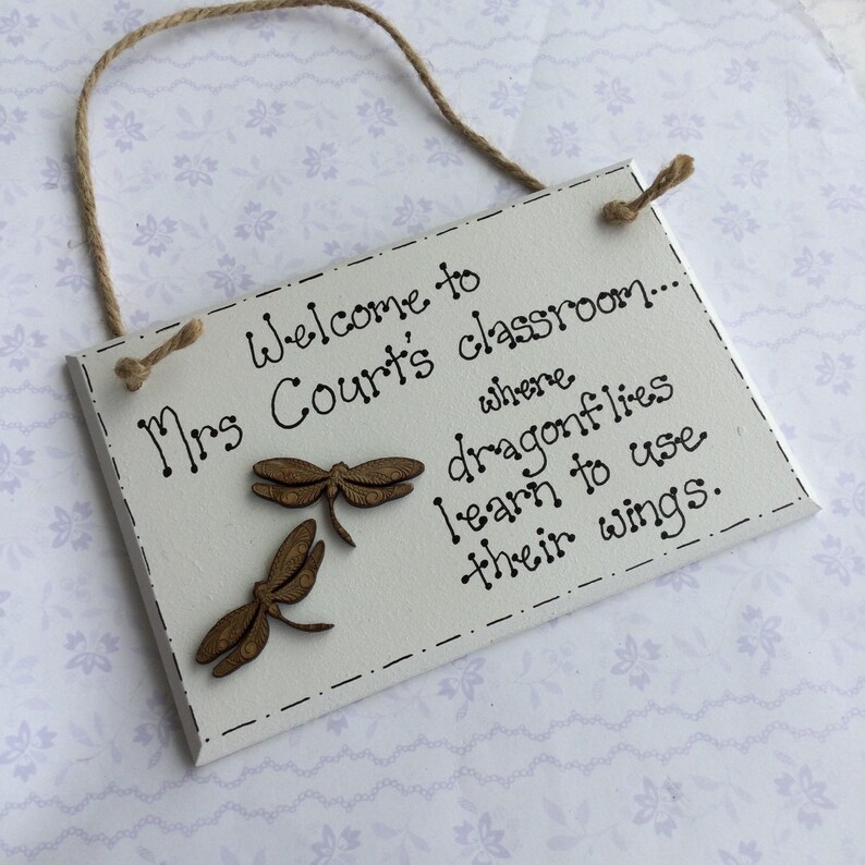 May include: White wooden sign with the text "Welcome to Mrs. Court's classroom... where dragonflies learn to use their wings." Two brown wooden dragonfly cutouts are attached to the sign.
