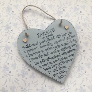 Puede incluir: Un letrero de madera en forma de coraz&oacute;n azul claro con el nombre "Kayleigh" y una descripci&oacute;n escrita en tinta negra. El letrero tiene dos peque&ntilde;os corazones de madera y un lazo de cuerda para colgar.