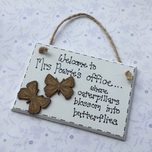 May include: White wooden sign with the text "Welcome to Mrs. Powrie's office... where caterpillars blossom into butterflies". The sign has two brown wooden butterfly decorations.