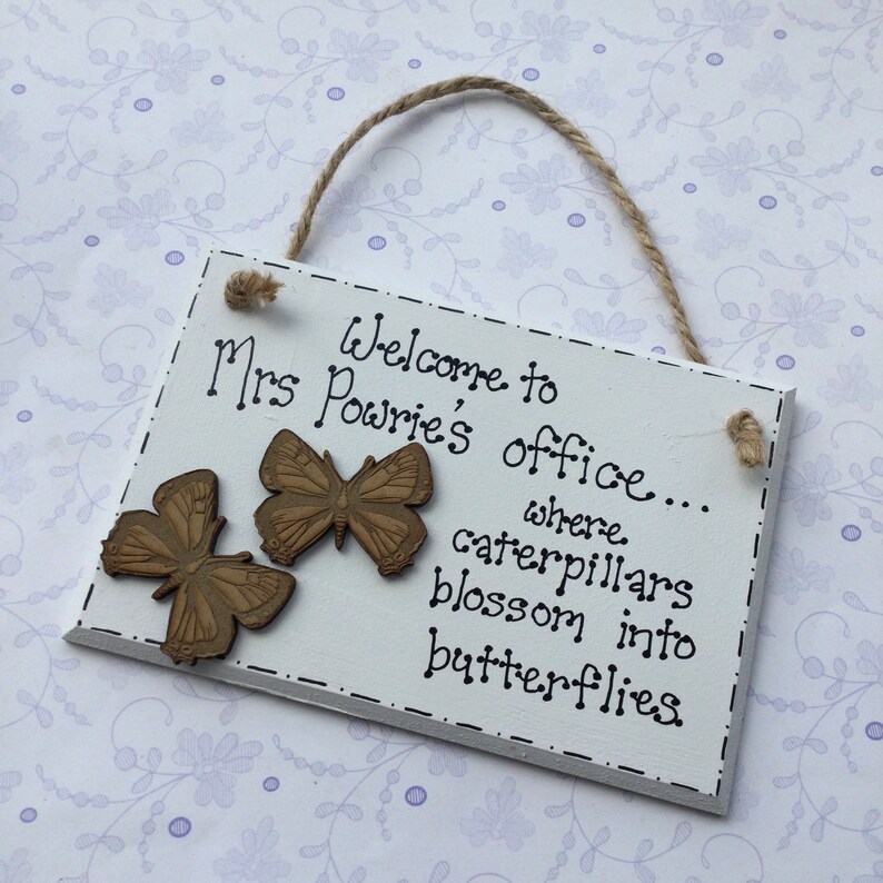 May include: White wooden sign with the text "Welcome to Mrs. Powrie's office... where caterpillars blossom into butterflies". The sign has two brown wooden butterfly decorations.