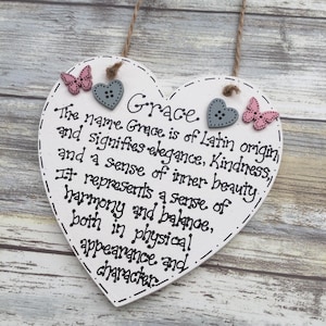 Puede incluir: Un letrero de madera en forma de coraz&oacute;n blanco con el nombre "Grace" y su significado en texto negro. Decorado con botones de coraz&oacute;n grises y acentos de mariposa rosas. El letrero est&aacute; suspendido por una cuerda natural. Medidas no aplicables.