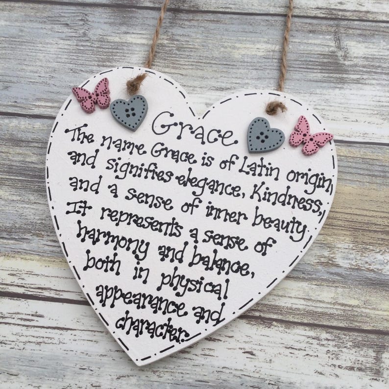 Puede incluir: Un letrero de madera en forma de coraz&oacute;n blanco con el nombre "Grace" y su significado en texto negro. Decorado con botones de coraz&oacute;n grises y acentos de mariposa rosas. El letrero est&aacute; suspendido por una cuerda natural. Medidas no aplicables.