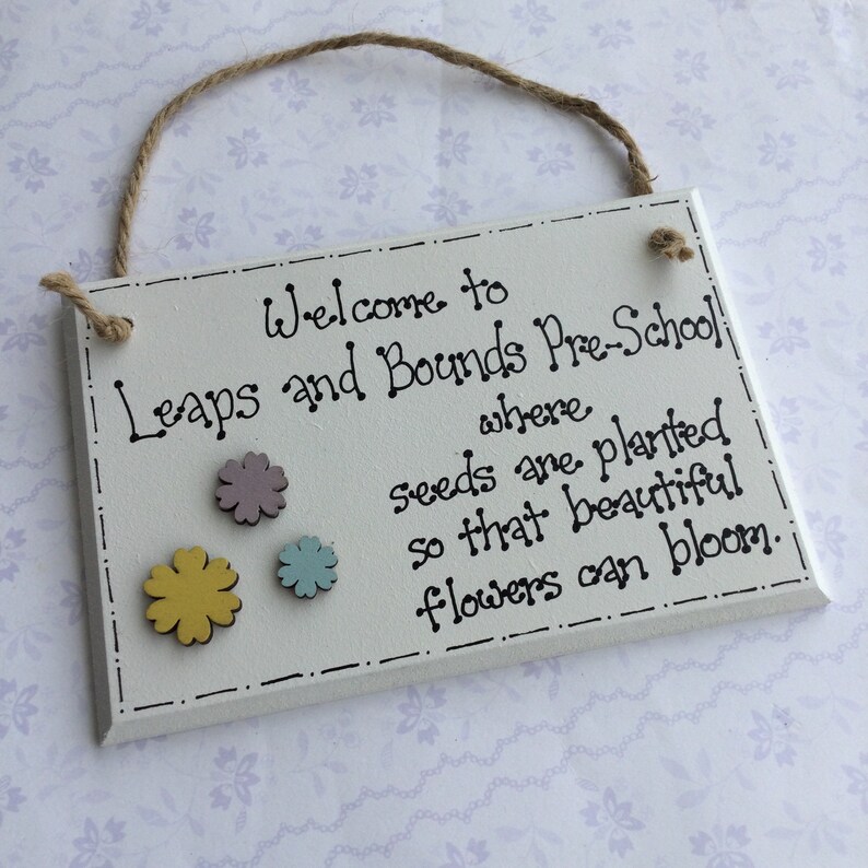 May include: White wooden sign with black text that reads "Welcome to Leaps and Bounds Pre-School where seeds are planted so that beautiful flowers can bloom." The sign has three small wooden flowers attached to it: one yellow, one light blue, and one light purple.