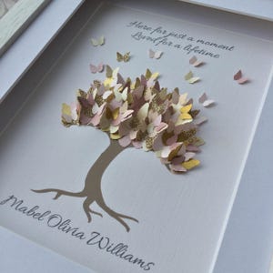May include: A framed artwork featuring a tree with a brown trunk and branches, adorned with paper butterflies in shades of pink, gold, and cream. The artwork includes the text "Here for just a moment, Loved for a lifetime" and a name.