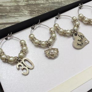 May include: A set of silver-toned wine glass charms featuring white faux pearls and clear rhinestones. Each hoop includes a unique dangling charm: a silver number 30, a silver seashell with a pearl center, and a silver heart engraved with K&G.