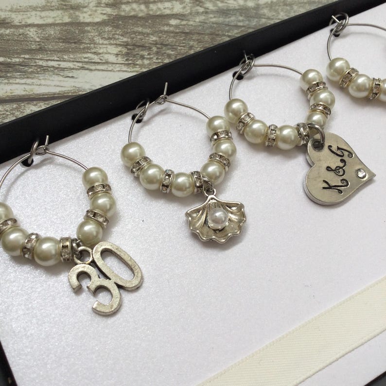 May include: A set of silver-toned wine glass charms featuring white faux pearls and clear rhinestones. Each hoop includes a unique dangling charm: a silver number 30, a silver seashell with a pearl center, and a silver heart engraved with K&G.