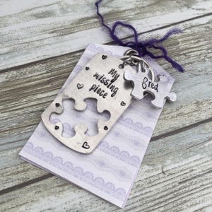 Może przedstawiać: Srebrny zestaw breloków w kształcie puzzli. Jeden element zawiera napis "My missing piece" z małymi akcentami w kształcie serca. Drugi element jest wygrawerowany imieniem "Fred". Zestaw jest przymocowany do fioletowego sznurka i spoczywa na lawendowej karcie.