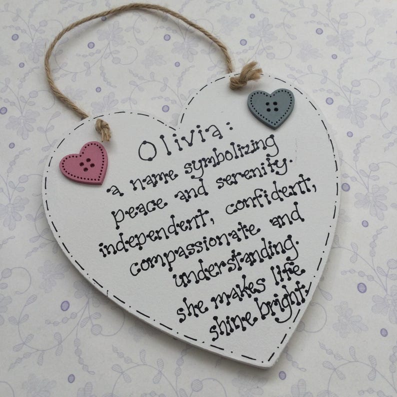 Puede incluir: Un letrero de madera en forma de coraz&oacute;n blanco con el nombre "Olivia" y una descripci&oacute;n de sus cualidades. Cuenta con dos botones en forma de coraz&oacute;n y un colgador de cuerda. El texto es negro.