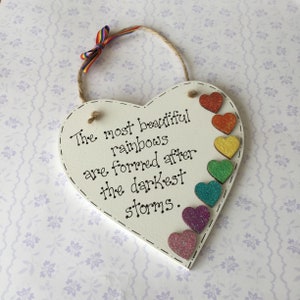 May include: White wooden heart-shaped sign with the text "The most beautiful rainbows are formed after the darkest storms."  The sign is decorated with a rainbow of glitter hearts.