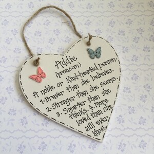 Puede incluir: Un letrero de madera en forma de coraz&oacute;n color crema con el nombre "Addie" y una lista de atributos positivos en texto negro. Cuenta con dos adornos de mariposa y una cuerda de yute para colgar. Los bordes est&aacute;n decorados con una l&iacute;nea punteada.