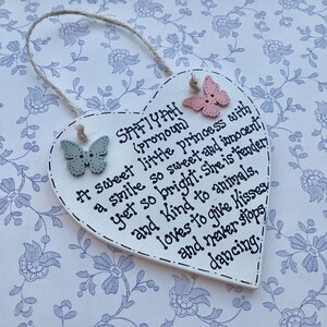 Puede incluir: Placa de madera en forma de coraz&oacute;n con el nombre "SAFIYAH" y un poema escrito con tinta negra. La placa tiene dos adornos en forma de mariposa y un colgador de cuerda. El fondo es un estampado floral.