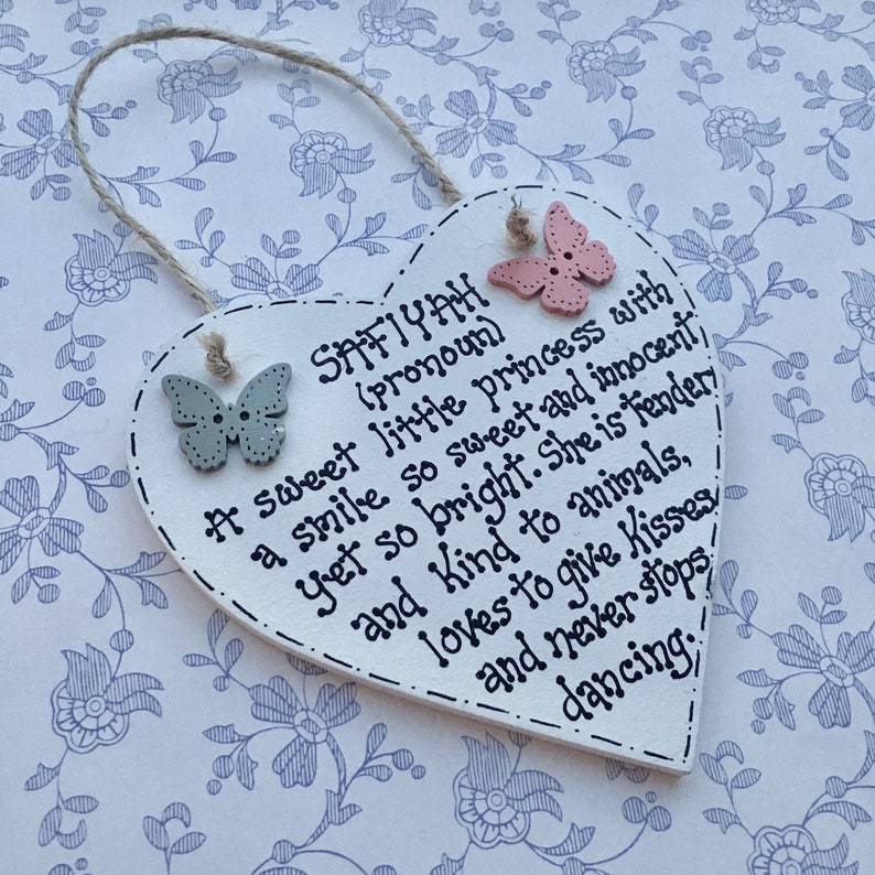 Puede incluir: Placa de madera en forma de coraz&oacute;n con el nombre "SAFIYAH" y un poema escrito con tinta negra. La placa tiene dos adornos en forma de mariposa y un colgador de cuerda. El fondo es un estampado floral.