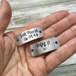 May include: Two rectangular silver-toned metal tags with rounded corners and two holes on each side. One tag is stamped with the text Just Married and the date 26.09.26. The other tag is stamped with the text Wifey H and a small heart symbol.