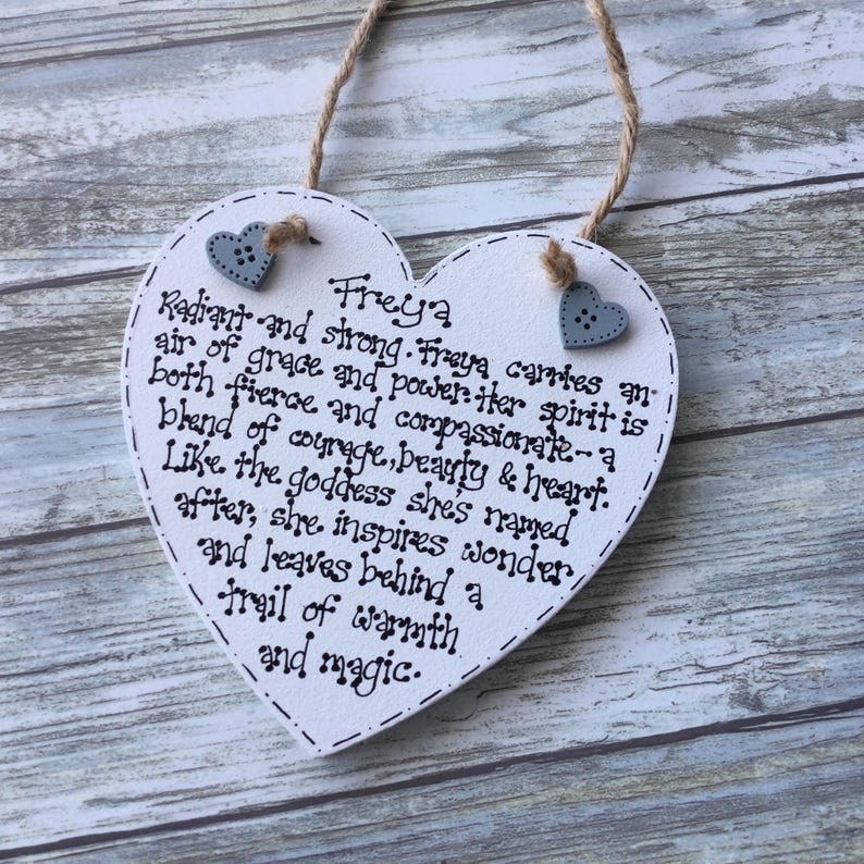 Puede incluir: Una placa de madera blanca en forma de coraz&oacute;n con el nombre "Freya" y un poema escrito con tinta negra. Se adjuntan dos peque&ntilde;os botones grises en forma de coraz&oacute;n. Un lazo de cuerda en la parte superior para colgar.